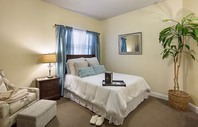 A cozy bedroom with a neatly made bed featuring white bedding and decorative pillows. There is a wooden nightstand with a lamp and a pair of glasses on it next to the bed. A cushioned armchair with a folded blanket and an ottoman are positioned near the bed. A large potted plant is in the corner, and a rectangular mirror hangs on the wall above the bed. Light blue curtains frame a window behind the bed.