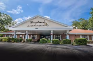 Front exterior view of Braybrook Assisted Living Facility showing a single-story building with a covered entrance, bushes, and a clear blue sky with some clouds.