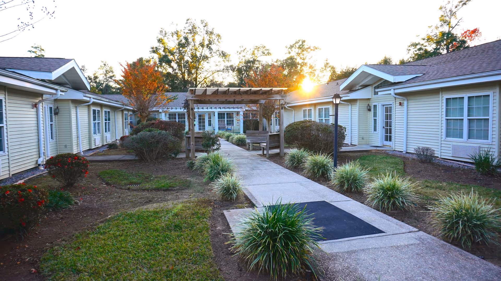 Outdoor courtyard area at a senior living facility with a concrete walkway lined by ornamental grasses and bushes. There is a wooden pergola with a bench underneath, surrounded by single-story buildings with light-colored siding and multiple windows. The sun is setting behind the trees in the background.