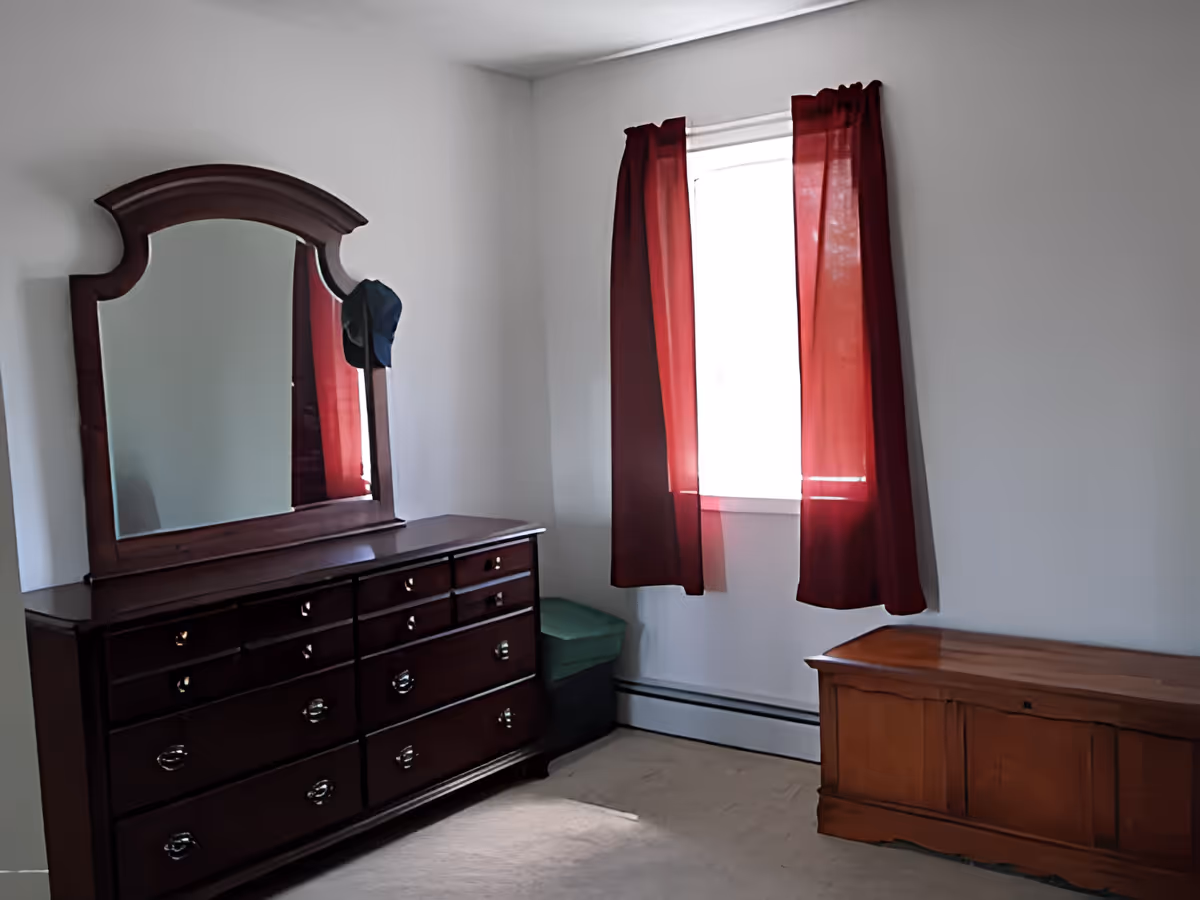 A simple bedroom corner with a dark wooden dresser featuring a large mirror, a window with red curtains letting in light, a green storage bin, and a wooden chest on a beige carpeted floor.