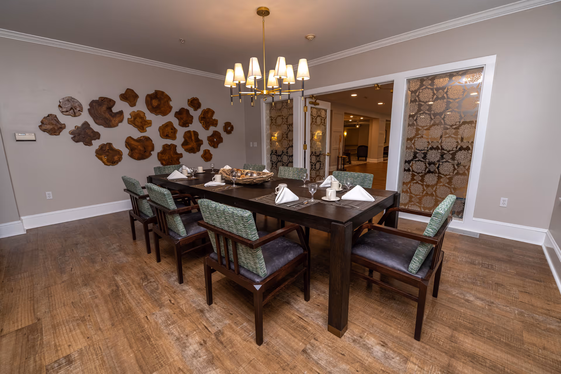 A dining room with a dark wooden table set for six people with white napkins, cups, and glasses. The chairs have wooden frames with green and black cushions. A modern chandelier with multiple lampshades hangs above the table. The room has wooden flooring, light-colored walls, decorative wooden wall art, and glass doors with patterned designs leading to another room.