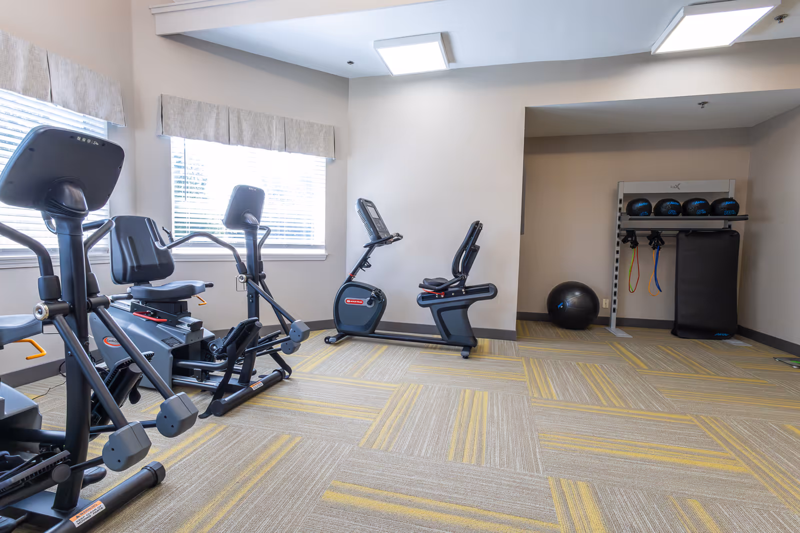 A small fitness room with exercise equipment including stationary bikes and a rowing machine. There is a rack with medicine balls, resistance bands, and a large exercise ball in the corner. The room has large windows with blinds and light-colored walls and carpet with yellow and gray patterns.