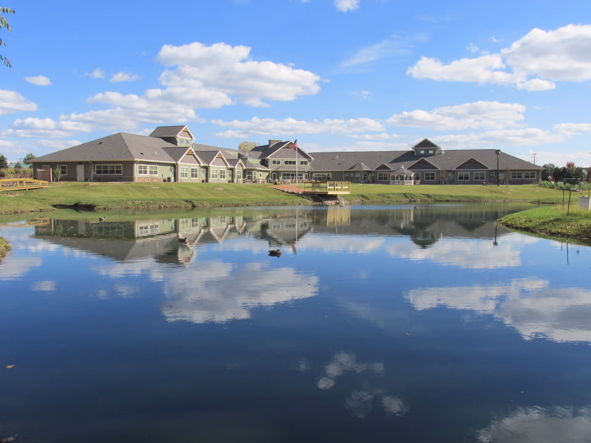 A large single-story assisted living facility building with multiple peaked roofs reflected in a calm pond in the foreground under a blue sky with scattered clouds.