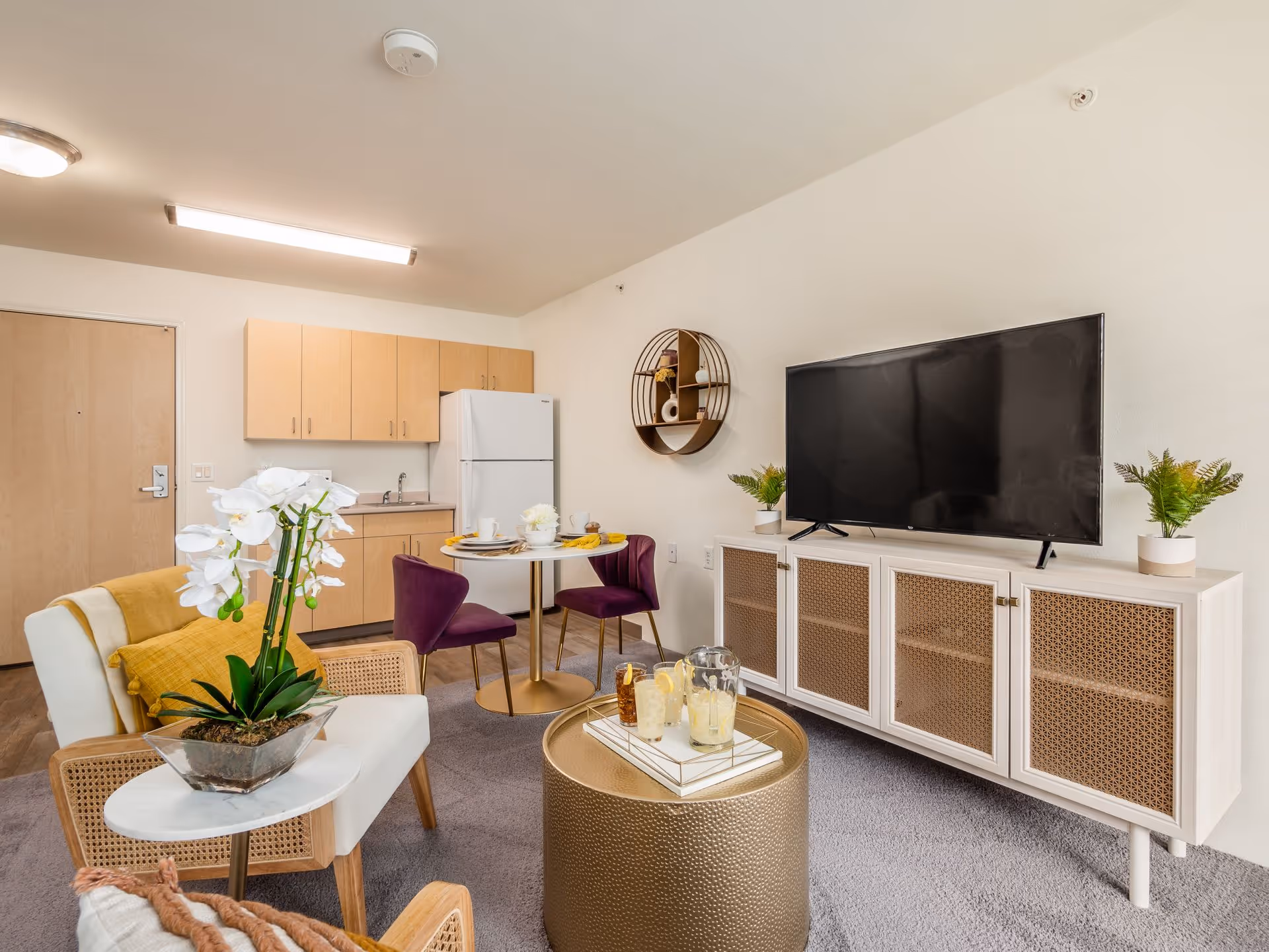 A cozy living area in a senior living facility featuring a small kitchenette with light wood cabinets and a white refrigerator. The room includes a round dining table with two purple chairs, a white armchair with a yellow throw blanket, a round gold coffee table with drinks, and a large flat-screen TV on a white cabinet with woven panel doors. Decorative plants and a circular wall shelf add to the warm and inviting atmosphere.