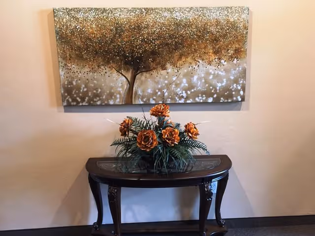 A decorative wooden console table with a glass inset top, holding a floral arrangement of orange flowers and green leaves, positioned against a beige wall. Above the table hangs a large rectangular painting depicting a tree with a dense, textured canopy in shades of brown, gold, and white.