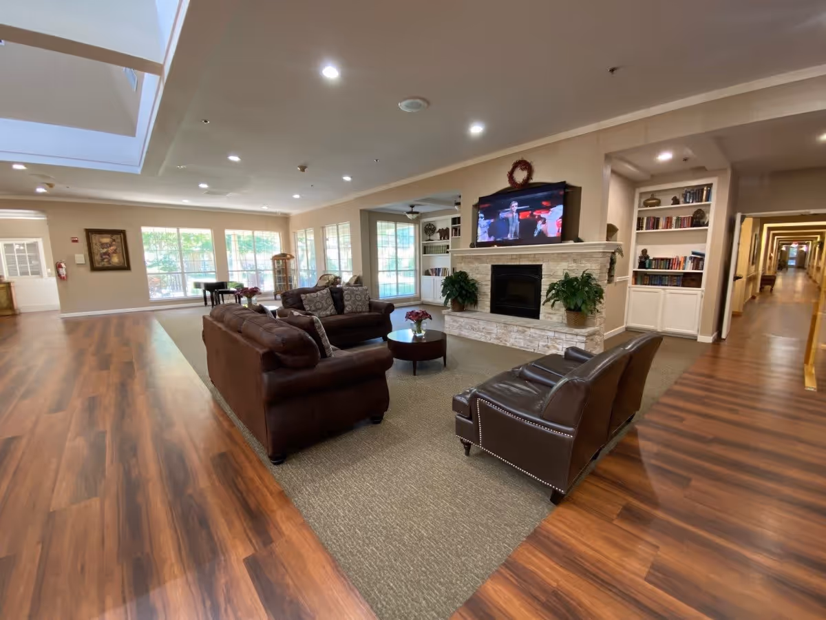 A spacious living room area in The Waterford on Cooper featuring brown leather sofas and armchairs arranged around a round coffee table. A stone fireplace with a TV mounted above it is centered on one wall, flanked by two potted plants. Built-in bookshelves with books and decorative items are visible to the right. Large windows in the background let in natural light, and a long hallway extends to the right. The floor is a combination of wood and carpet.