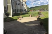 Outdoor paved patio area with several potted plants arranged along the edges, adjacent to a multi-story building with white and beige siding. Green grass surrounds the patio, and there is a hill and partly cloudy sky in the background.