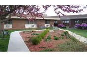 Outdoor courtyard area at Spurgeon Manor featuring a curved concrete walkway, landscaped garden beds with small shrubs and mulch, a bench under a tree with pink blossoms, and a brick building with windows in the background.