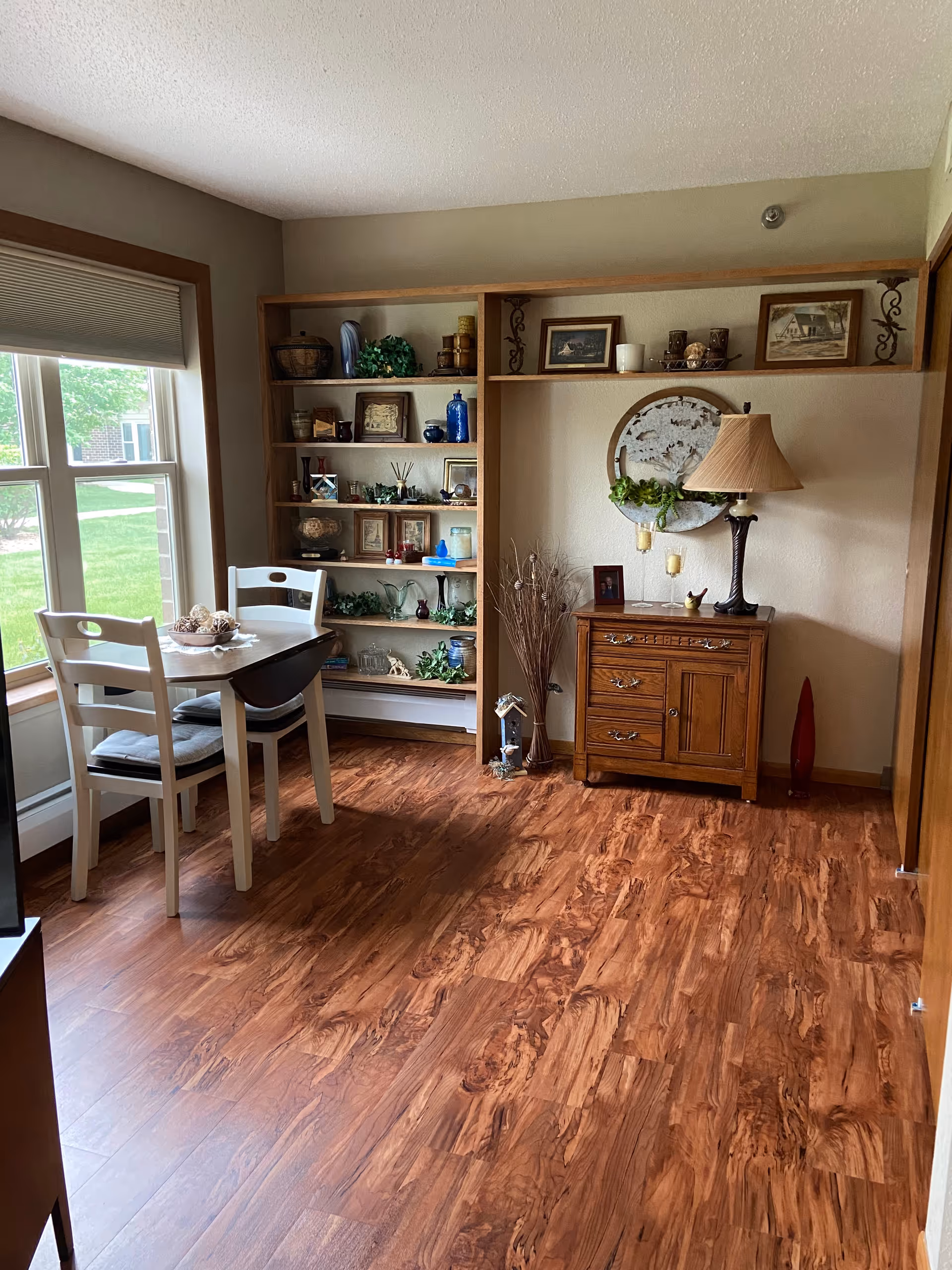 A cozy interior room with wooden flooring, a small dining table with two white chairs near a large window, and a wooden shelving unit filled with decorative items and framed pictures. There is also a wooden cabinet with a table lamp, candles, and a decorative wall hanging above it.