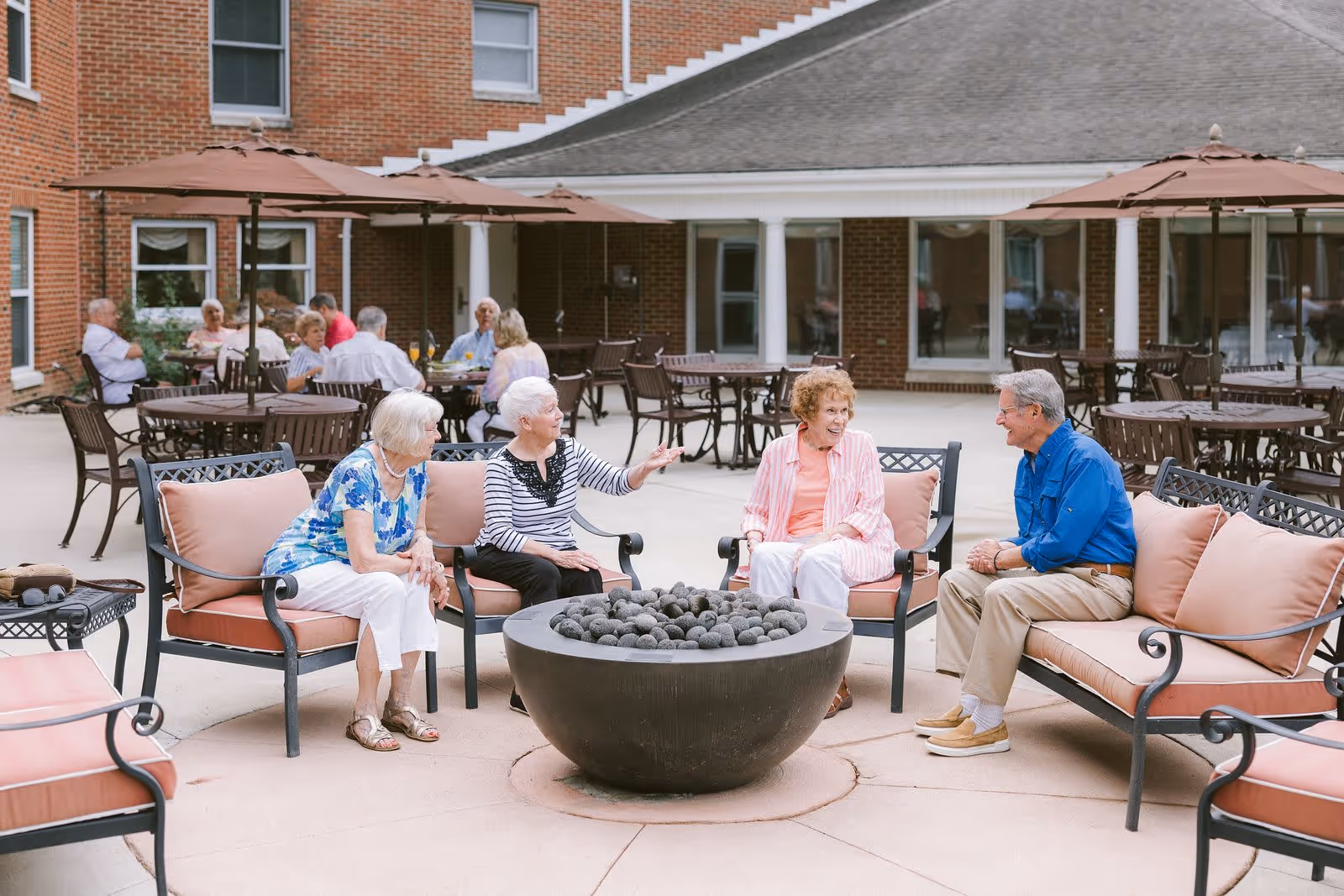 A group of elderly people sitting and chatting around a circular fire pit in an outdoor patio area of a senior living facility. The patio has cushioned metal chairs and tables with umbrellas, and a brick building with windows and white columns is visible in the background.
