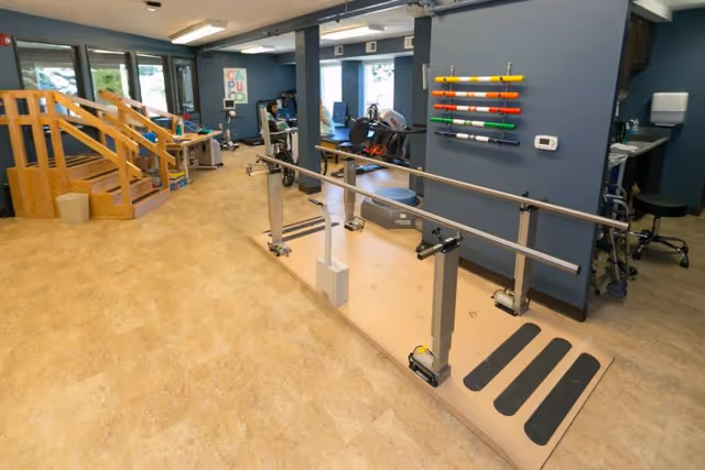 Physical therapy room with parallel walking bars, practice stairs, exercise machines and colorful therapy rods mounted on a blue wall.