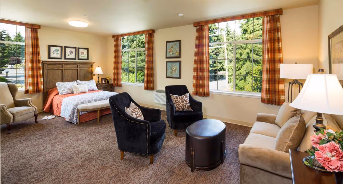 A spacious and well-lit bedroom in a senior living facility with a bed featuring a wooden headboard, two black armchairs with patterned pillows, a beige sofa, a small round ottoman, and large windows with plaid curtains showing green trees outside. The room also has a beige armchair, bedside table with a lamp, framed floral artwork on the walls, and a vase with pink flowers on a side table.