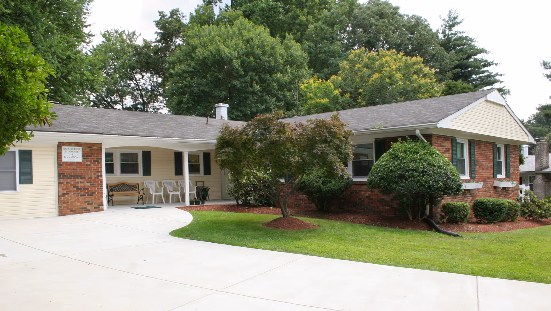 Exterior view of a single-story senior living facility named AlfredHouse II, featuring a brick and beige siding facade, a covered entrance with chairs and a bench, surrounded by green grass, shrubs, and trees.