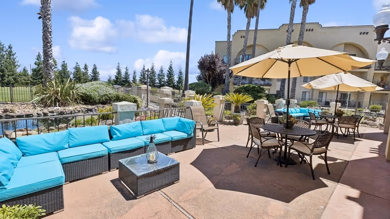 Outdoor patio area at Rio Las Palmas featuring turquoise cushioned wicker seating, several tables with beige umbrellas, surrounded by palm trees and landscaped greenery under a blue sky with scattered clouds.