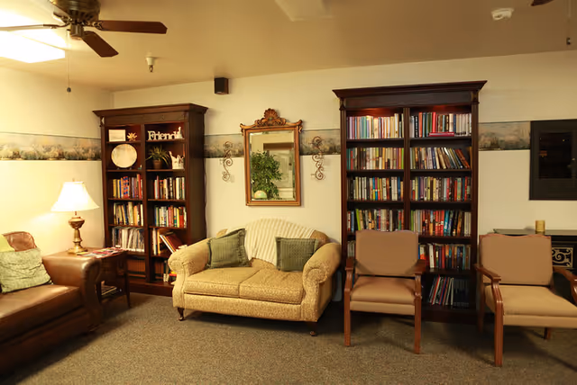Cozy common room with a loveseat, armchairs, two tall bookshelves, a table lamp, and a mirror on the wall.