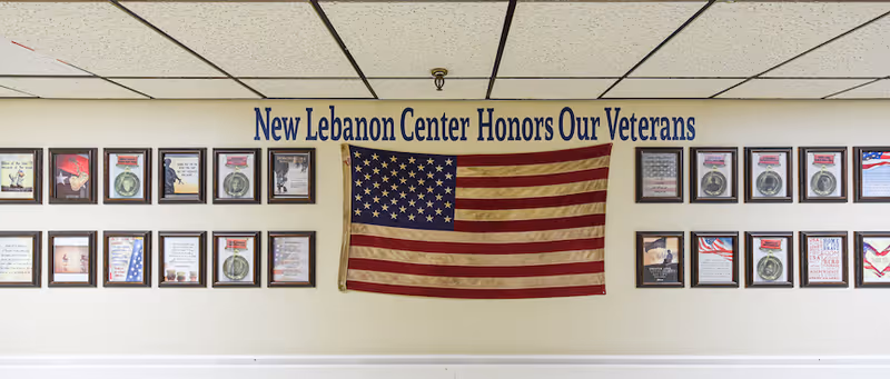 A hallway wall inside New Lebanon displaying an American flag, framed plaques, and the sign 'New Lebanon Center Honors Our Veterans'.