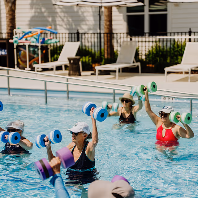 A group of older adults participating in a water aerobics class in an outdoor swimming pool, each holding colorful foam dumbbells above the water. Lounge chairs and poolside equipment are visible in the background.