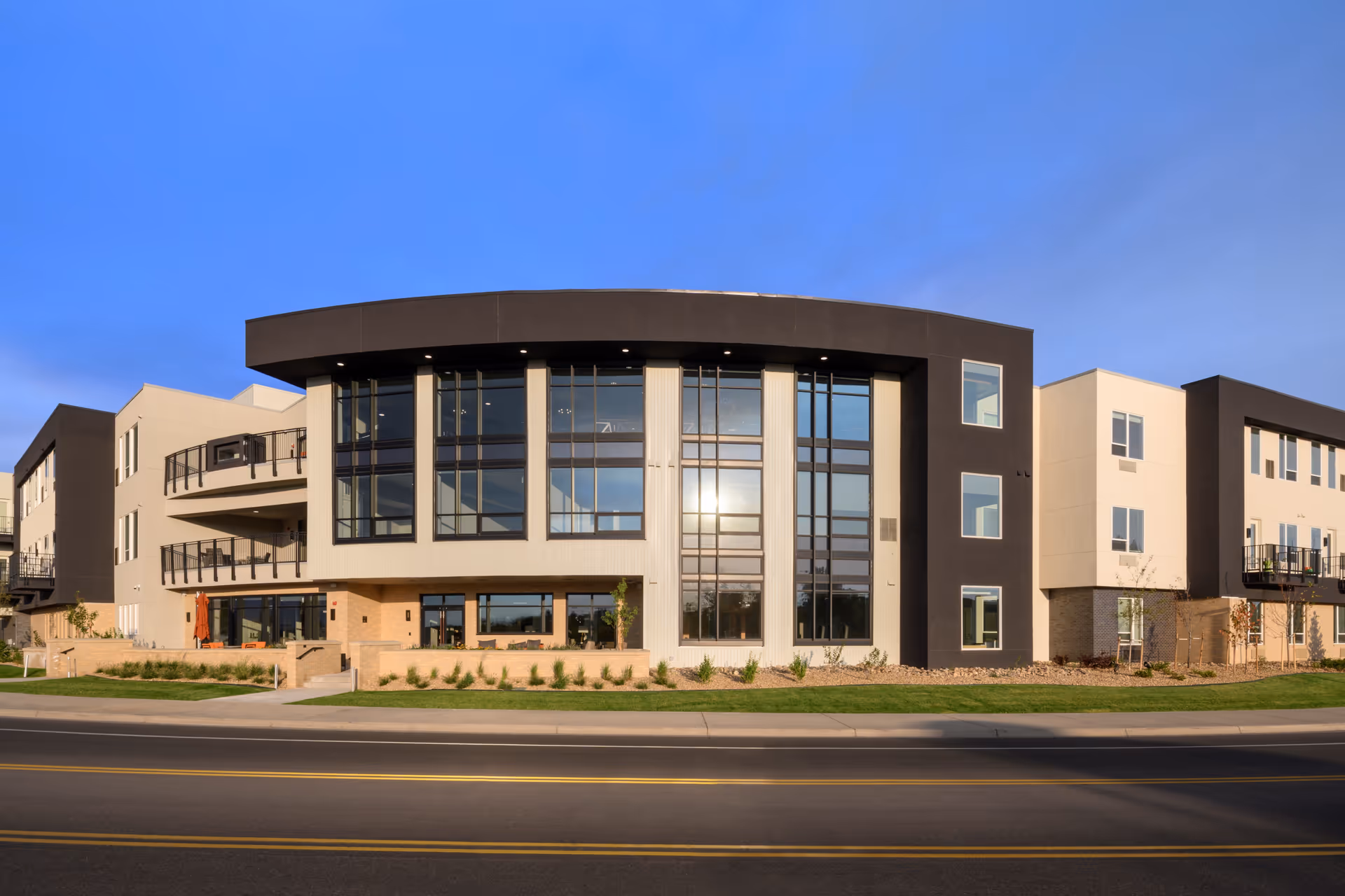 Exterior view of a modern three-story senior living facility building with large windows, balconies, and landscaped greenery in front, under a clear blue sky.