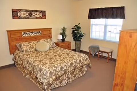 A bedroom with a double bed covered in a patterned bedspread, a wooden headboard, a nightstand with a lamp and plant, a large potted plant near a window with a dark valance, and a wooden dresser. There is also a small bench under the window.
