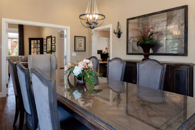 A dining room with a large glass-top wooden table surrounded by upholstered chairs. A floral centerpiece is placed on the table. The room features a chandelier hanging from the ceiling, a large decorative mirror on the wall, and a dark wooden sideboard with a floral arrangement. Doorways lead to adjacent rooms with additional furniture and decor visible.