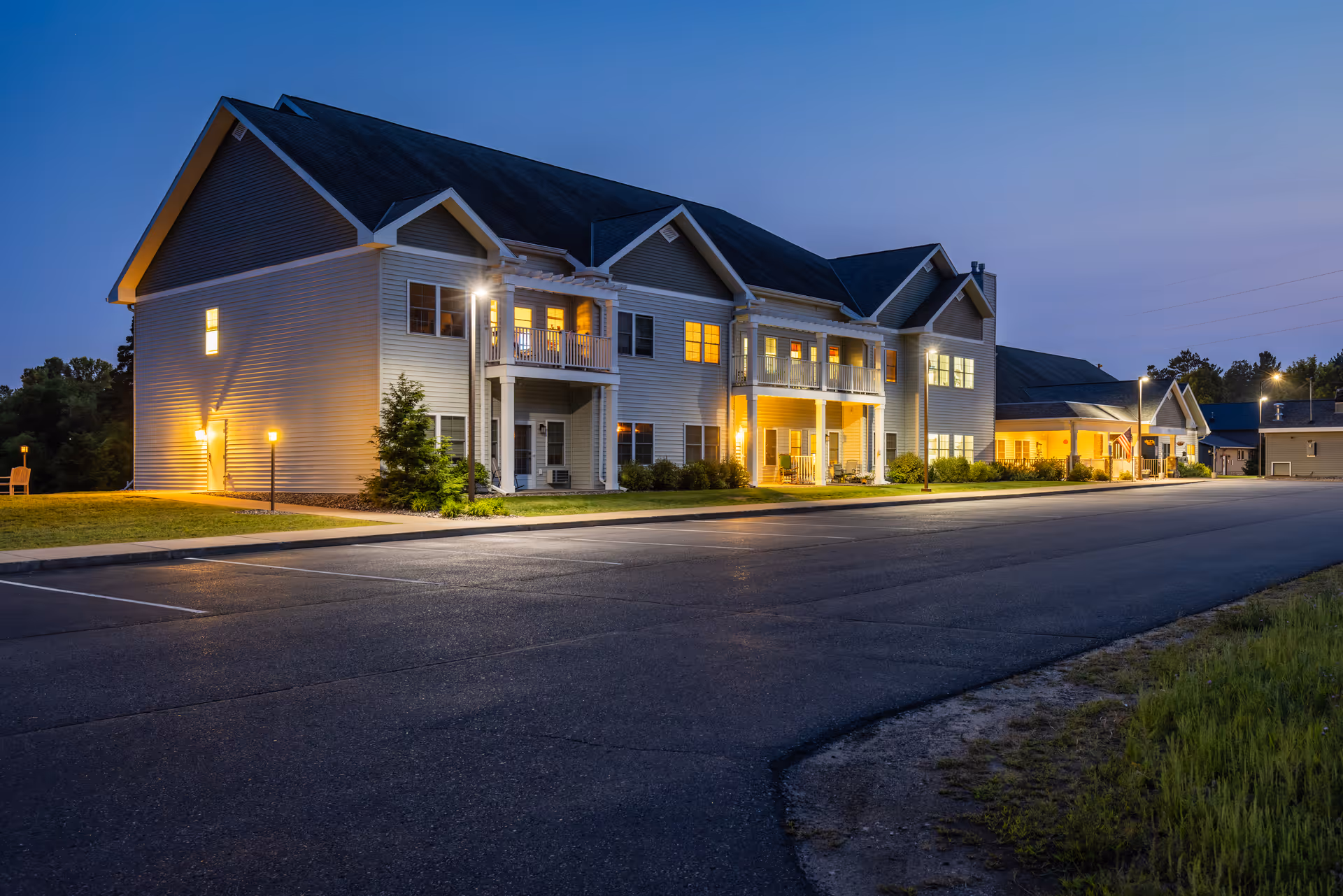 Exterior view of a two-story senior living facility building at dusk with lights on inside and outside. The building has white siding, multiple windows, balconies, and a paved parking area in front. The sky is clear and darkening, indicating early evening.