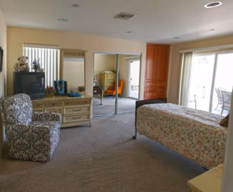 A furnished senior living bedroom with a bed, dresser, armchair, and mirrored closet.