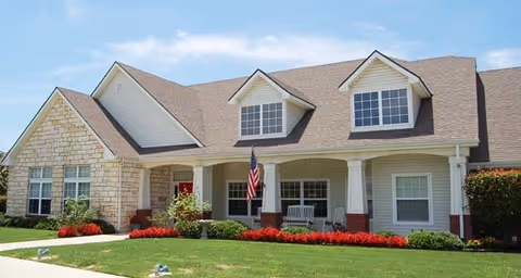 Front exterior view of a single-story residential building with a combination of stone and siding walls, a brown shingled roof with two dormer windows, a covered porch with white columns, an American flag, and well-maintained landscaping including green grass and red flowers.