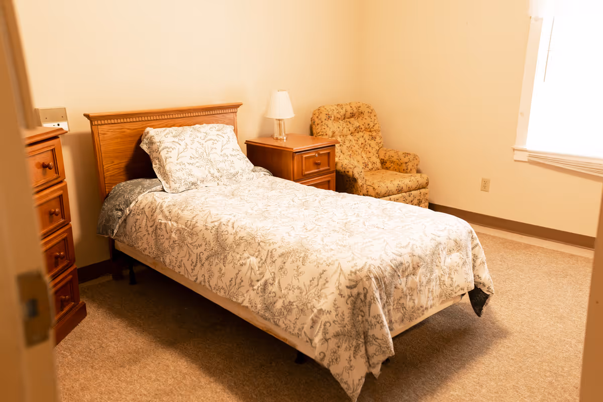 A small bedroom with a single bed dressed in patterned bedding, a wooden headboard and nightstand, and a floral upholstered armchair.