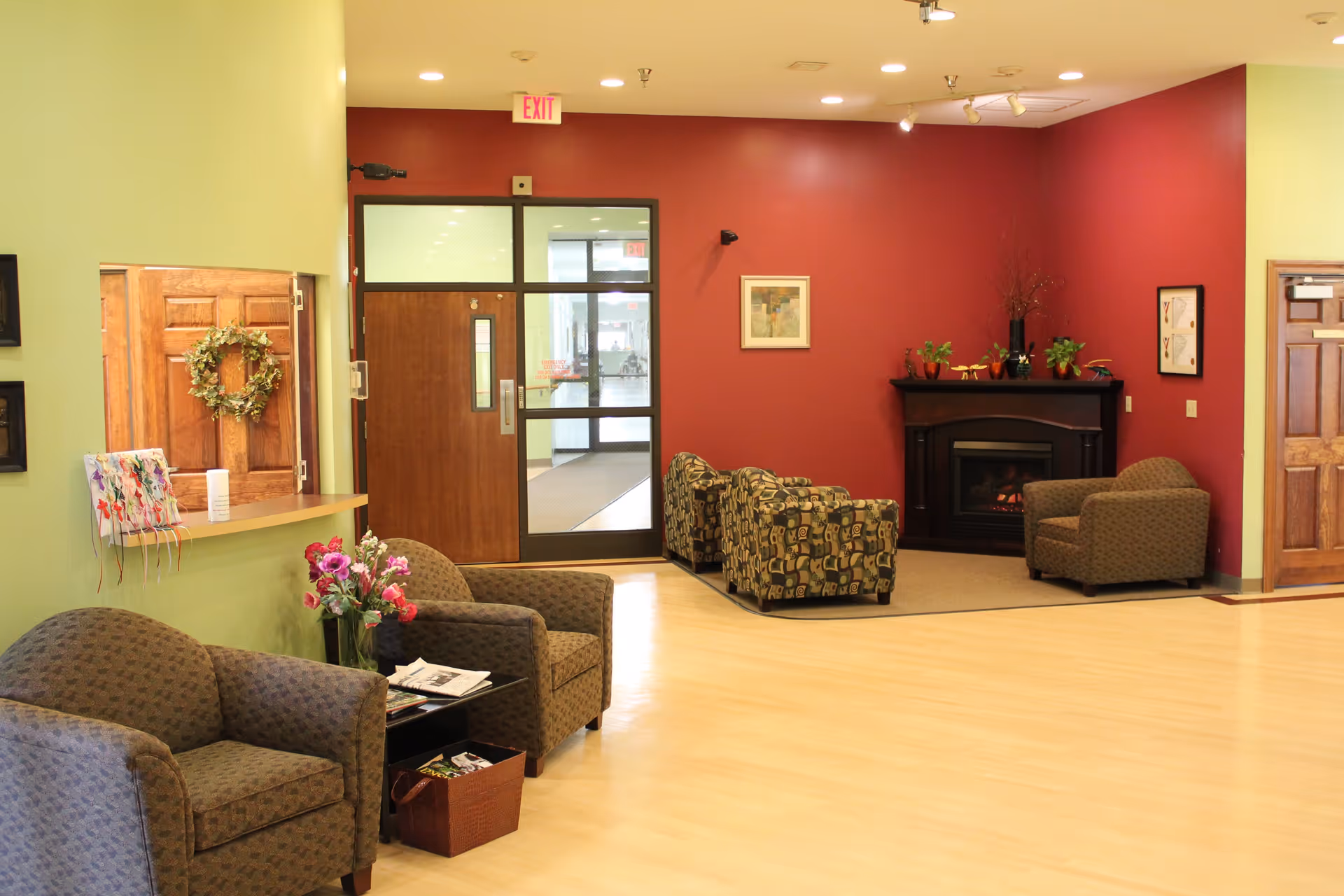 A cozy senior living facility common area with patterned armchairs arranged near a fireplace on a red accent wall. The room has light wood flooring, green walls, and wooden doors. There are framed pictures on the walls and a small table with flowers and magazines.