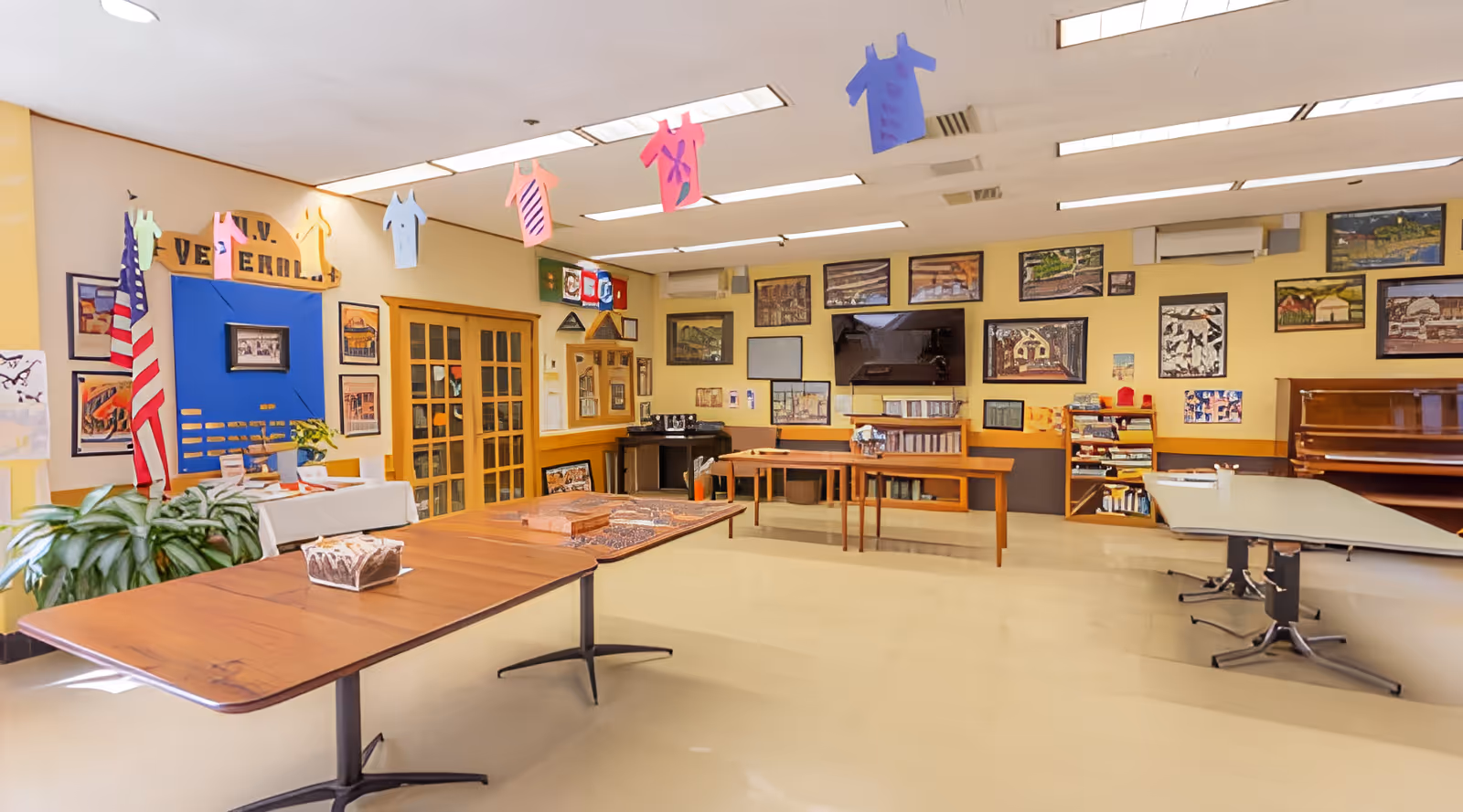 A bright activity room in a nursing facility with folding tables, a piano, wall-mounted TV and framed artwork, plus hanging paper shirts and an American flag.