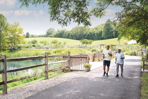 Two people walking along a paved path beside a pond and wooden fence in a landscaped outdoor area with rolling grassy hills and trees.