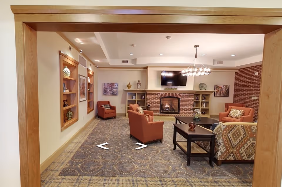 A cozy living room with a patterned carpet, several upholstered armchairs and a sofa arranged around a dark wooden coffee table. A brick fireplace with a mounted flat-screen TV above it is centered on the far wall, flanked by built-in shelves with decorative items. The room is softly lit by a modern chandelier and recessed ceiling lights. The walls are decorated with framed artwork and the space is viewed through a wooden door frame.