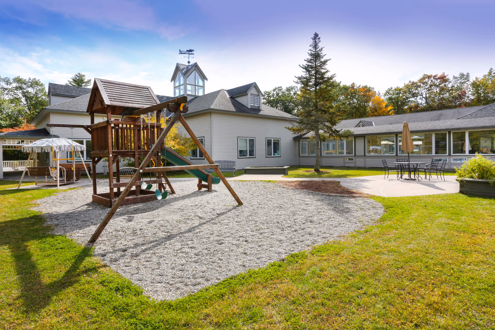Outdoor playground area with a wooden swing set and slide on a gravel surface, surrounded by green grass. In the background, there is a light gray building with multiple windows and a small tower with a weather vane on top. There is also a patio area with a table, chairs, and an umbrella. Trees with autumn foliage are visible behind the building under a partly cloudy sky.