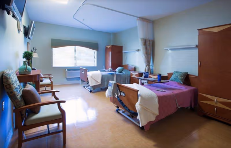 A bright room with two hospital-style beds, each with a blanket and pillow. There are wooden wardrobes and bedside tables next to each bed. Two chairs with cushions are placed near a small wooden table with a plant and a lamp. A large window with blinds lets in natural light, and an air conditioning unit is installed below the window.