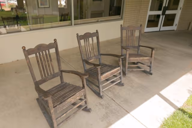 Three wooden rocking chairs placed on a concrete porch outside a building with large windows and double doors.