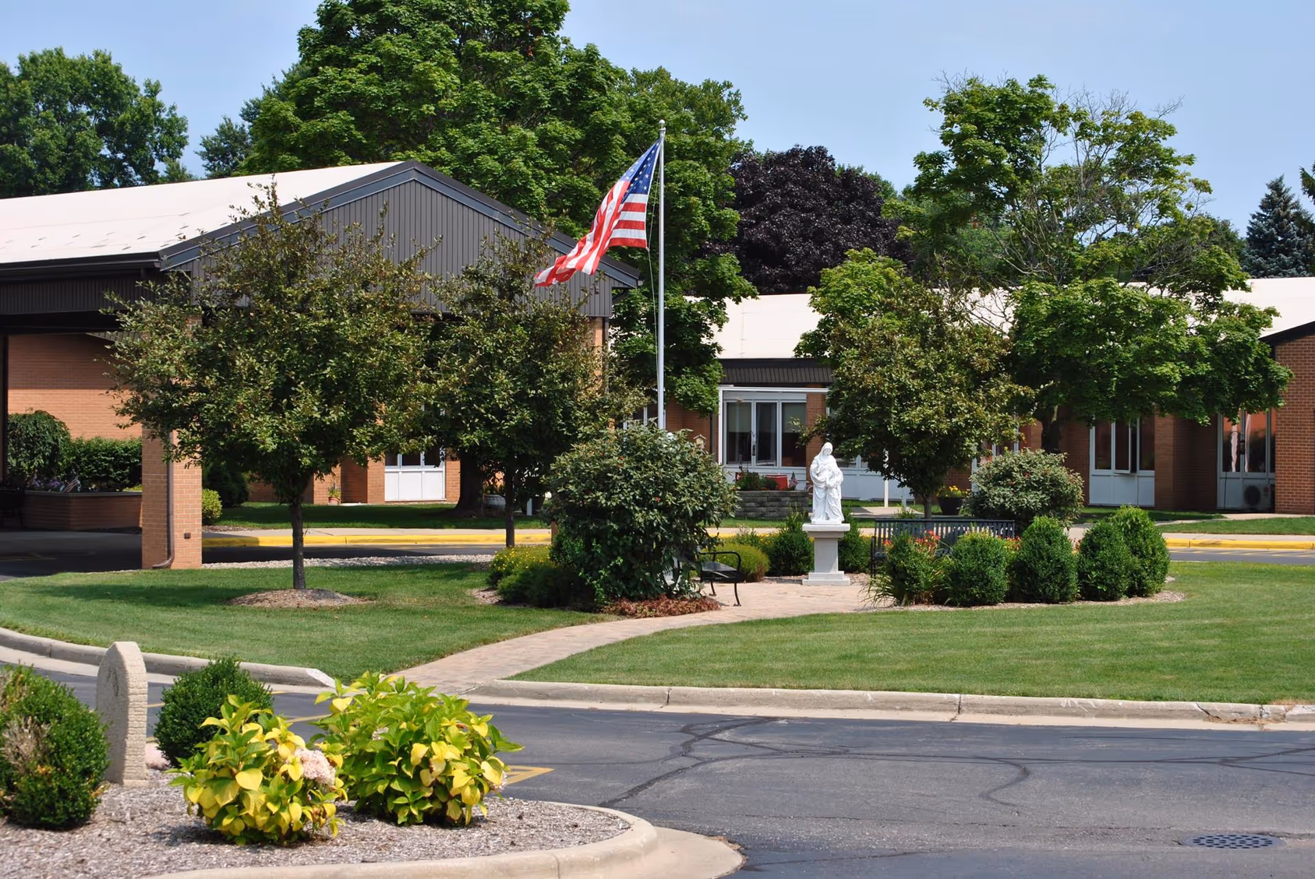 Front entrance of a brick senior living facility with a landscaped lawn, American flag, walkway, and a white statue near the driveway.