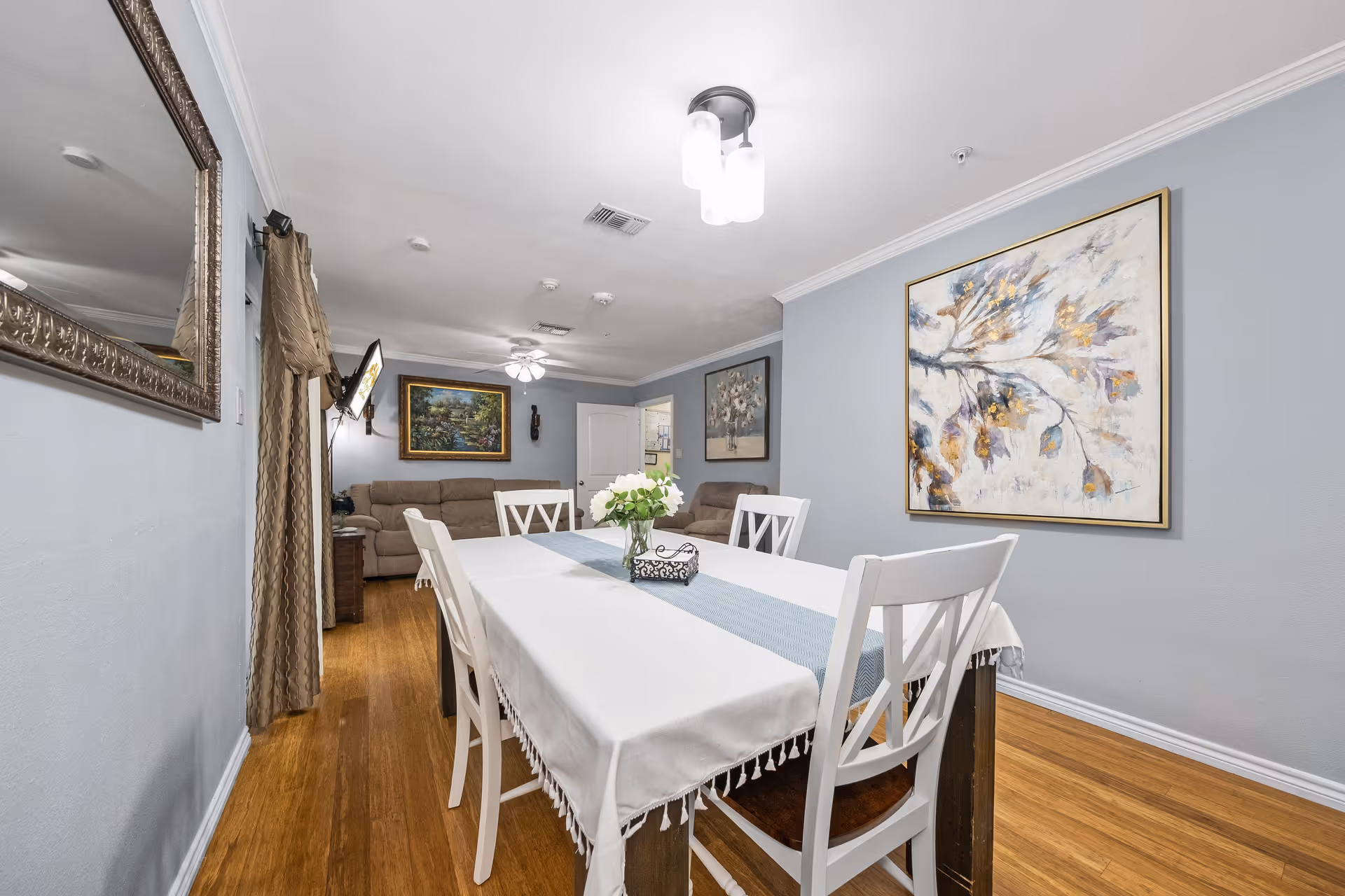 A dining area with a rectangular table covered with a white tablecloth and a blue runner, surrounded by white chairs. A vase with white flowers and a decorative tissue box are on the table. The room has light blue walls with framed artwork, wooden flooring, and a ceiling light fixture. In the background, there is a living area with beige sofas, a wall-mounted TV, and additional framed paintings.