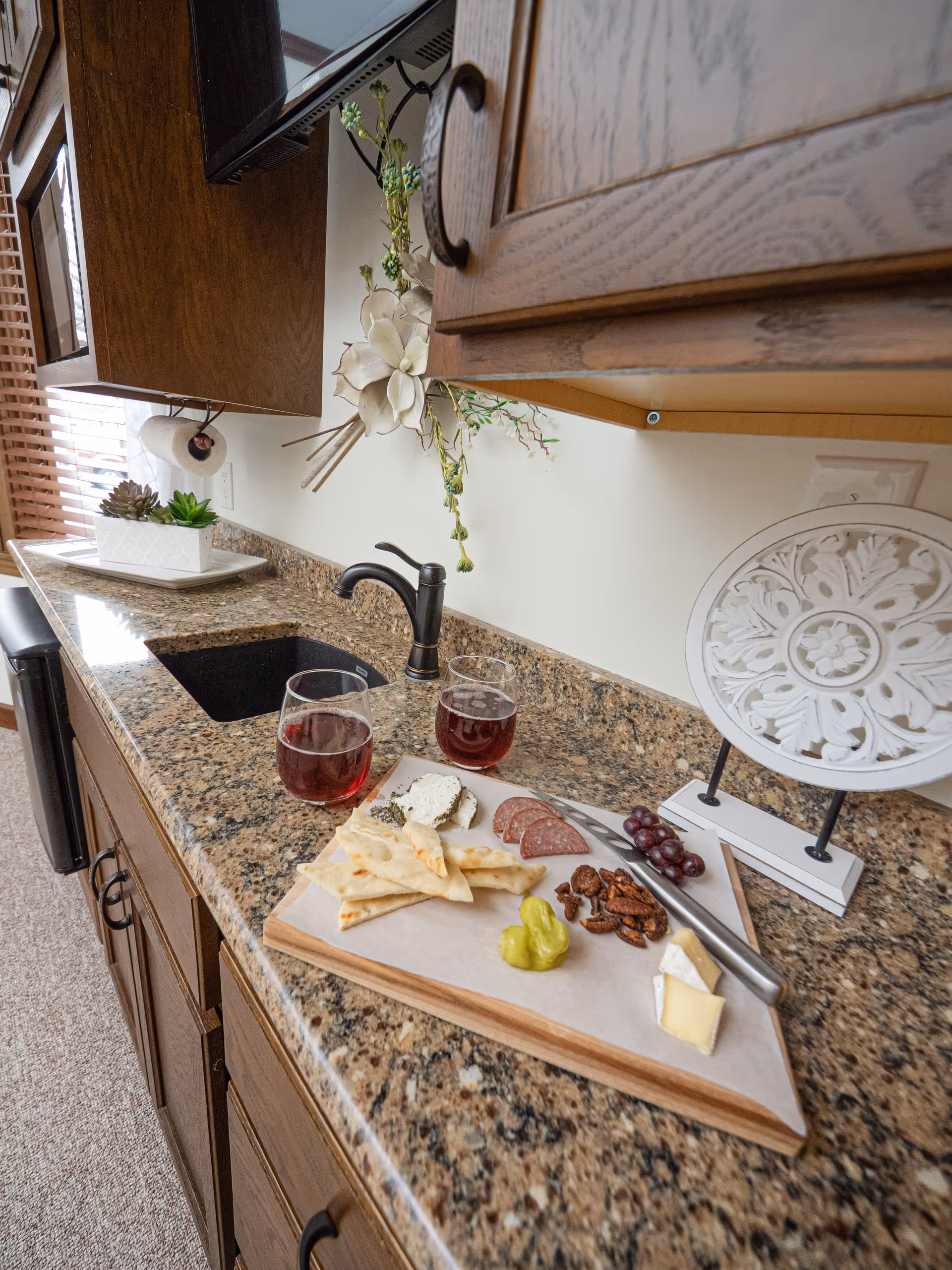 Granite kitchen countertop with a sink, wooden cabinets, a charcuterie board and two glasses of red wine.