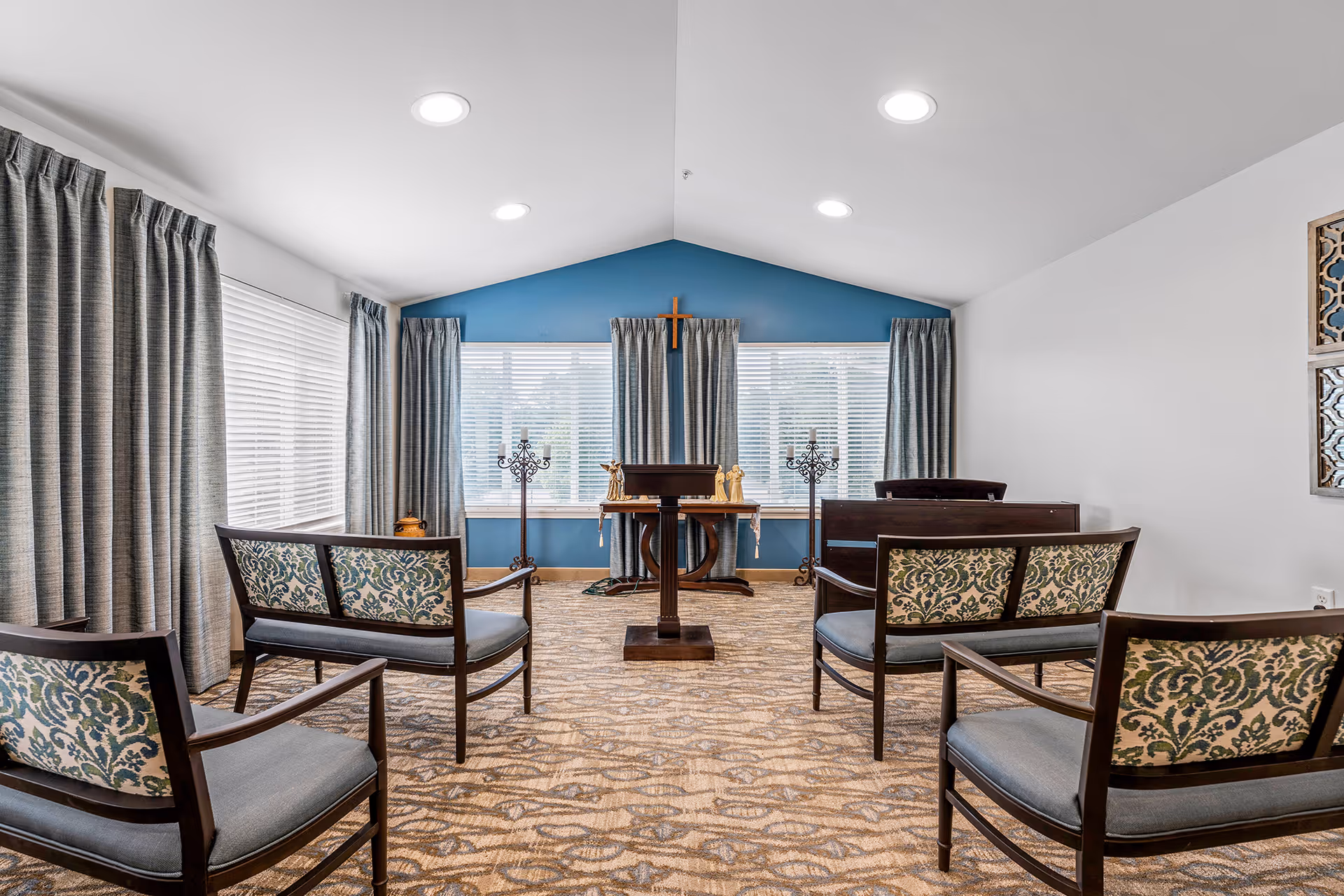 Small chapel-style meeting room with upholstered chairs facing a lectern and a cross on a blue accent wall.