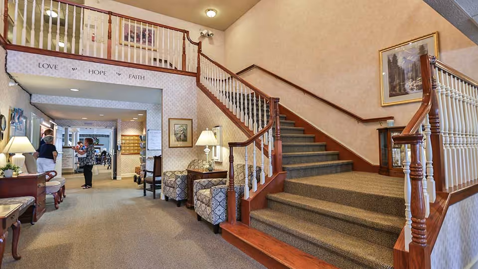 Interior view of a senior living facility lobby area with a carpeted staircase featuring wooden railings. The space includes patterned armchairs, side tables with lamps, framed artwork on the walls, and a hallway leading to other rooms where two people are conversing. Above the hallway entrance, the words 'LOVE', 'HOPE', and 'FAITH' are displayed on the wall.