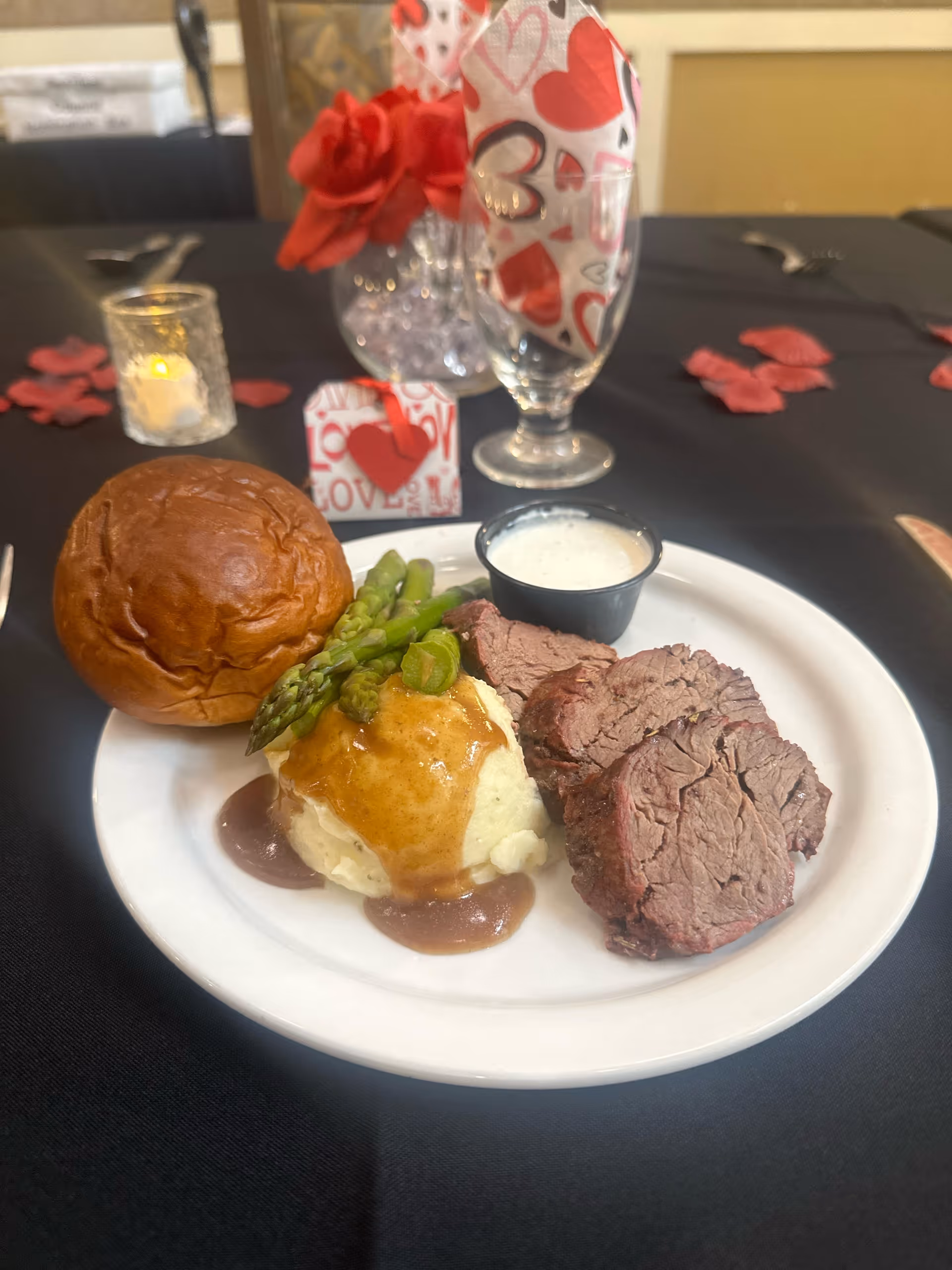 A plated meal consisting of three slices of roast beef, mashed potatoes with brown gravy, steamed asparagus, a bread roll, and a small cup of white sauce. The plate is on a black tablecloth with red rose petals scattered around. In the background, there is a glass with a heart-themed napkin, a small candle, and a small gift box with a red heart decoration.
