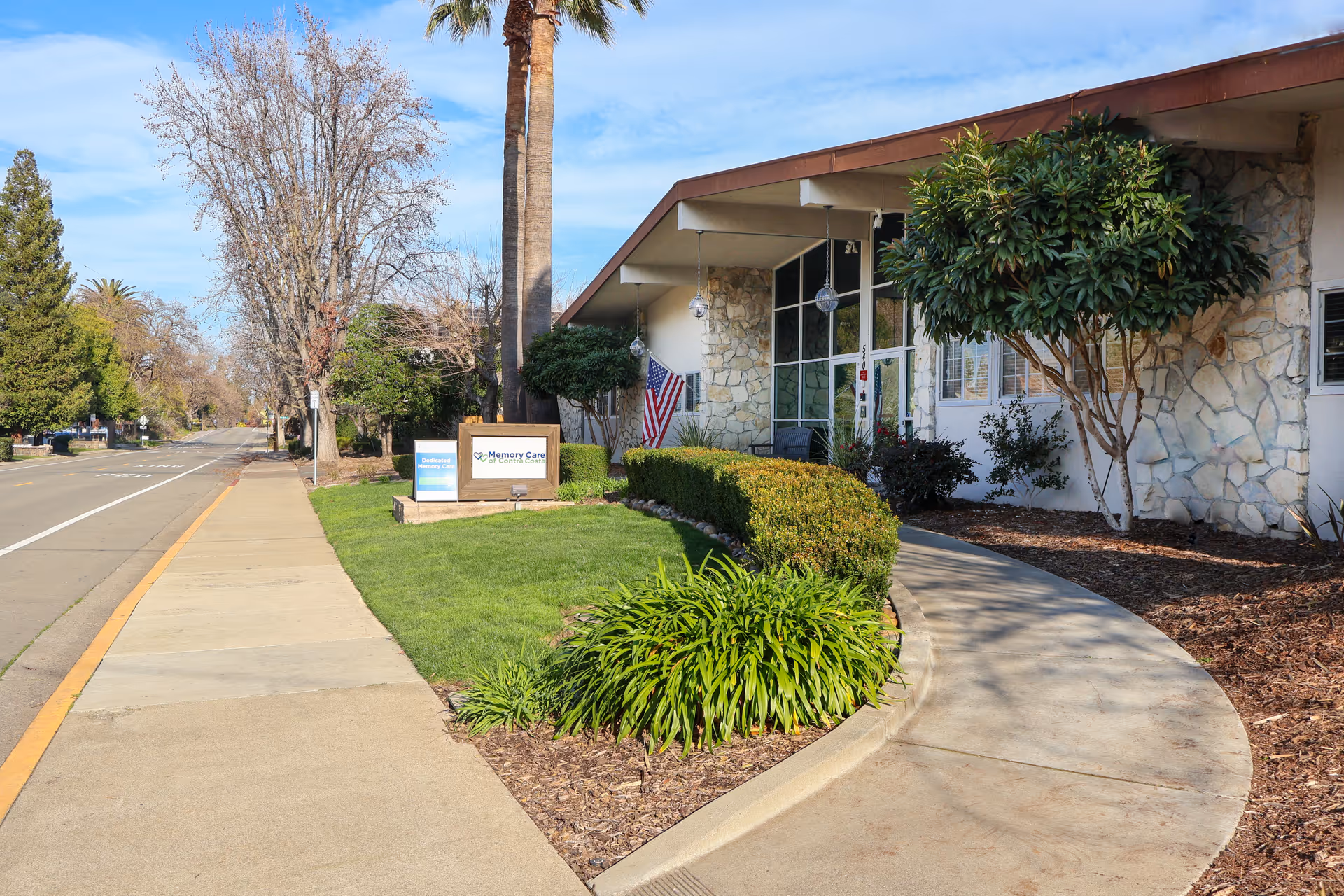 Exterior view of Memory Care of Contra Costa facility showing a stone facade building with large windows, a curved sidewalk, green bushes, and an American flag near the entrance. The scene includes a sidewalk along the street with trees and clear blue sky.