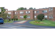 Exterior view of a brick apartment building with a driveway, cars, and neatly trimmed landscaping.