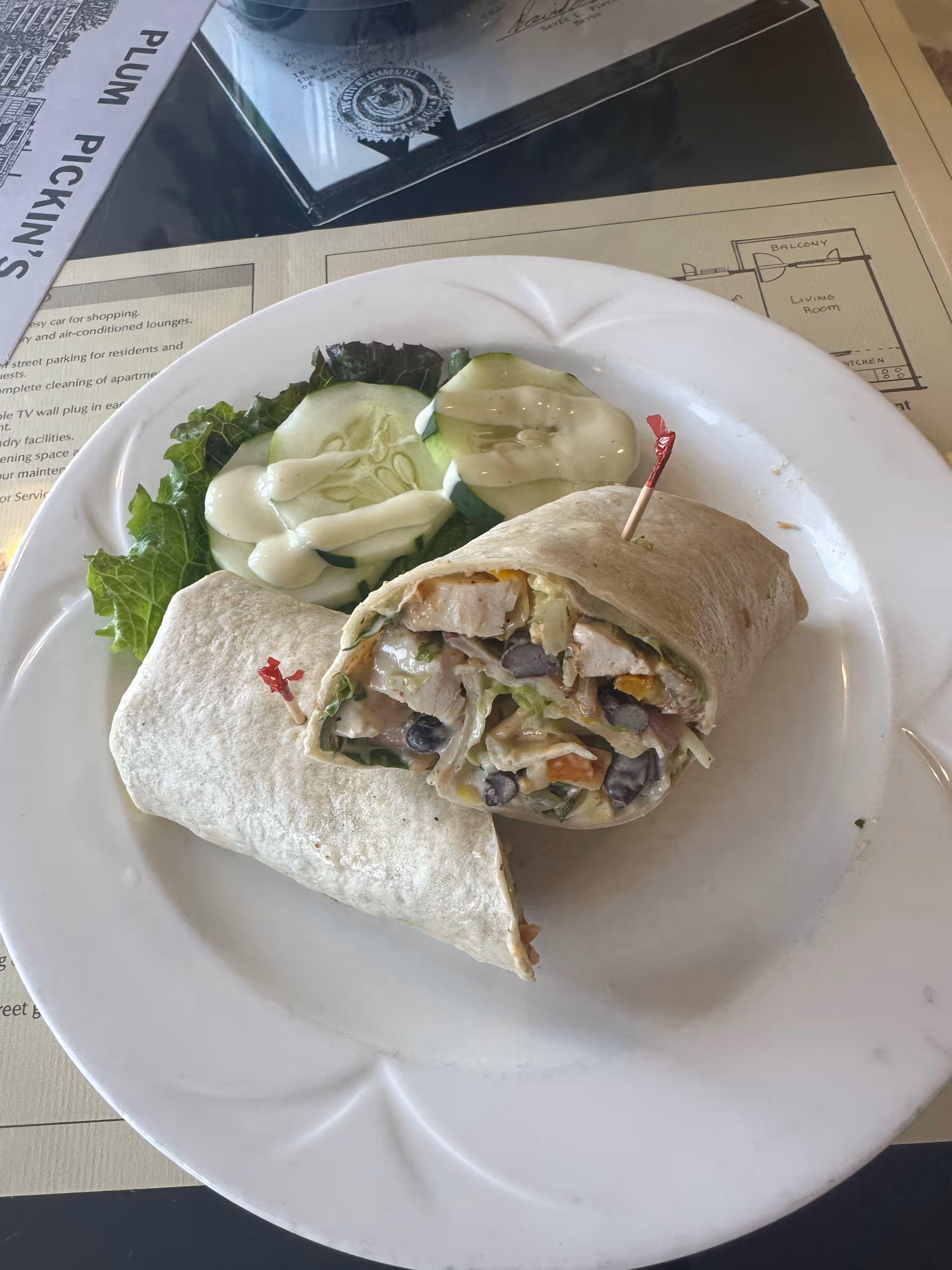 A white plate with a wrap sandwich cut in half, filled with chicken, black beans, lettuce, and other vegetables, accompanied by cucumber slices with dressing and a piece of lettuce. The plate is on a table with a placemat that has text and a floor plan.