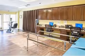 A spacious rehabilitation or therapy room with wooden flooring, featuring parallel bars for walking exercises, two computer workstations with chairs along a wall of wooden cabinets, and exercise equipment near large windows with natural light.