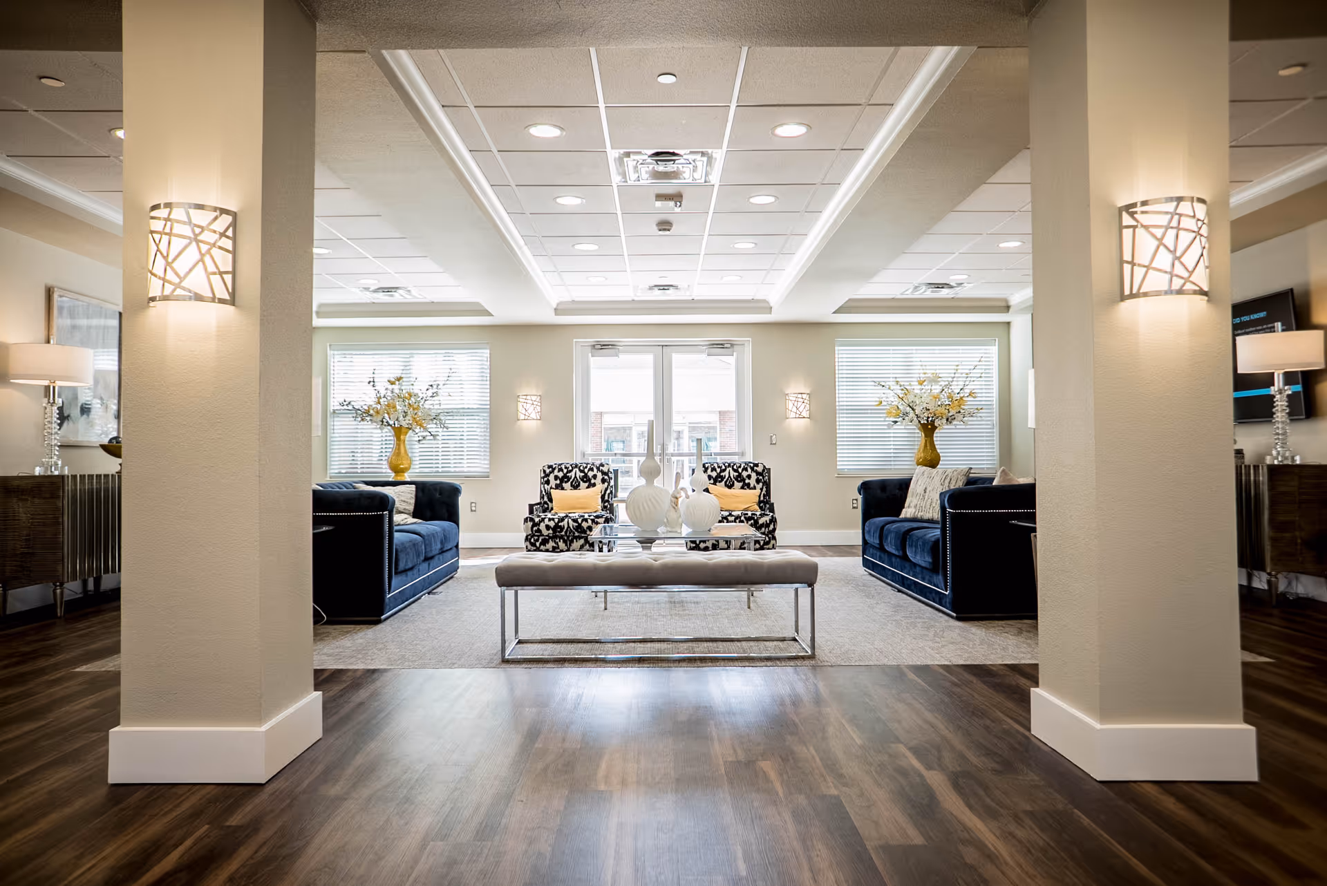 Bright, modern communal lounge with blue sofas, patterned armchairs, a bench-style coffee table and decorative vases between two pillars.