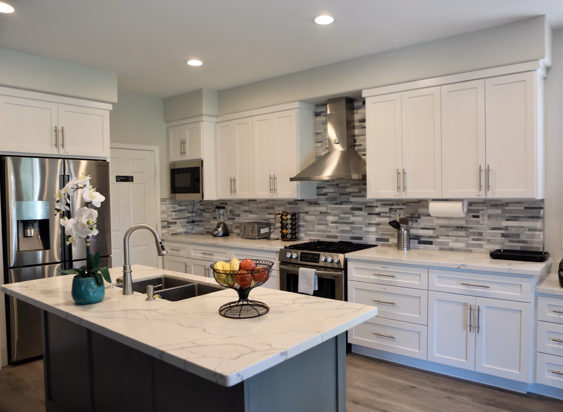 Modern kitchen with a marble-topped island, stainless steel appliances, white cabinets, and a tiled backsplash.