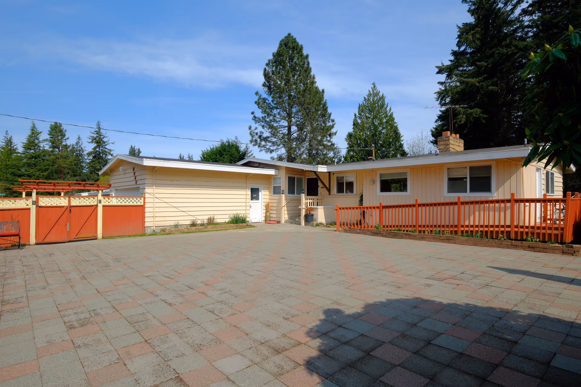 Exterior view of a single-story beige building with a red fence and gate, surrounded by tall trees under a blue sky with some clouds. The paved courtyard in front is spacious and clean.