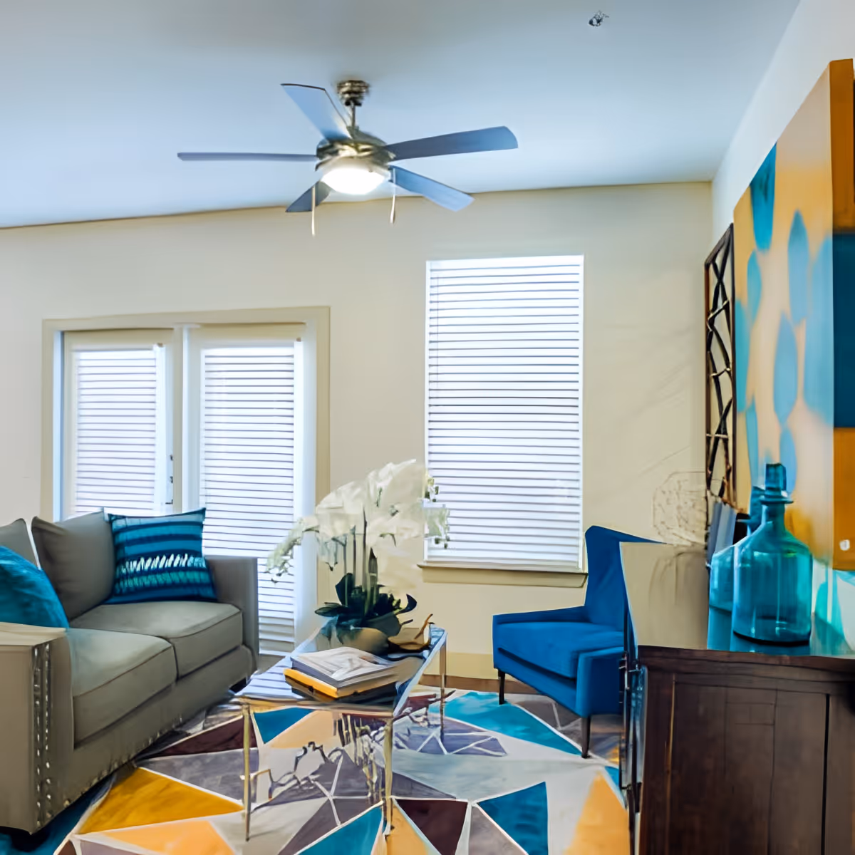 A modern living room with a gray sofa adorned with blue and teal pillows, a blue armchair, a glass coffee table with a white orchid plant and books, a colorful geometric patterned rug, a ceiling fan with light, and two windows with white blinds.