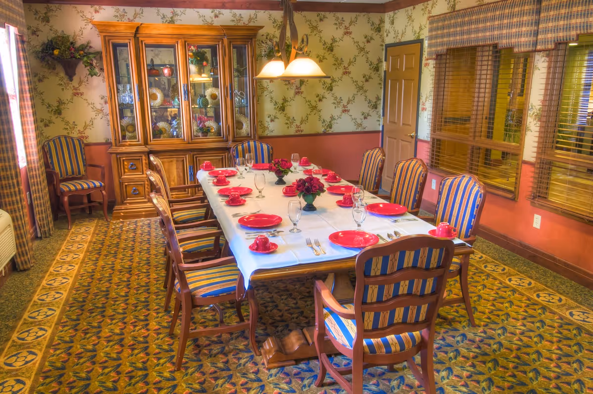 A dining room with a long rectangular table set for eight people. The table is covered with a white tablecloth and has red plates, cups, and saucers, along with glassware and silverware. There are three small floral centerpieces on the table. The chairs around the table have wooden frames with blue and yellow striped upholstery. The room has floral wallpaper, a patterned carpet, a wooden china cabinet filled with decorative items, and windows with wooden blinds.
